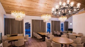 A modern dining area with wooden tables and chairs, large chandeliers, and large windows with curtains.