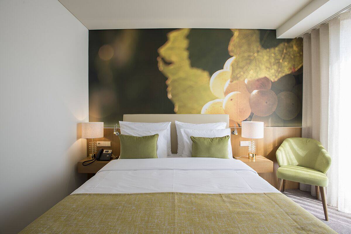 A hotel room with a large bed, two bedside lamps, a telephone, a green chair, and a wall mural.