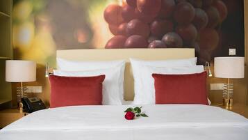 A hotel room with a large bed, red pillows, white bedding, a single red rose, and a wall with a grape cluster.