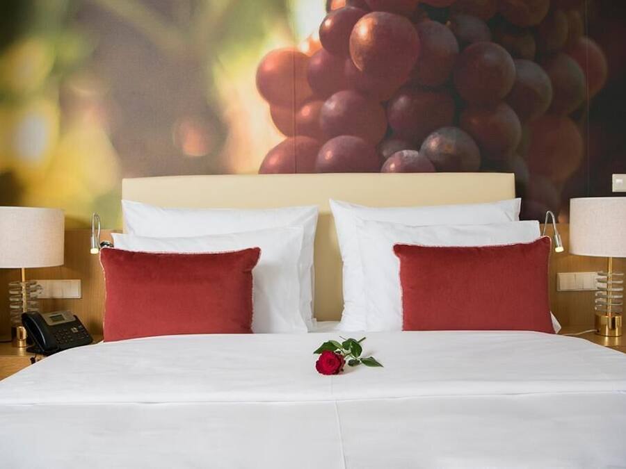 A hotel room with a large bed, red pillows, white bedding, a single red rose, and a wall with a grape cluster.