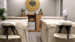 A room with two massage tables covered in beige cloths, a wooden cabinet with a mirror, and a small refrigerator.