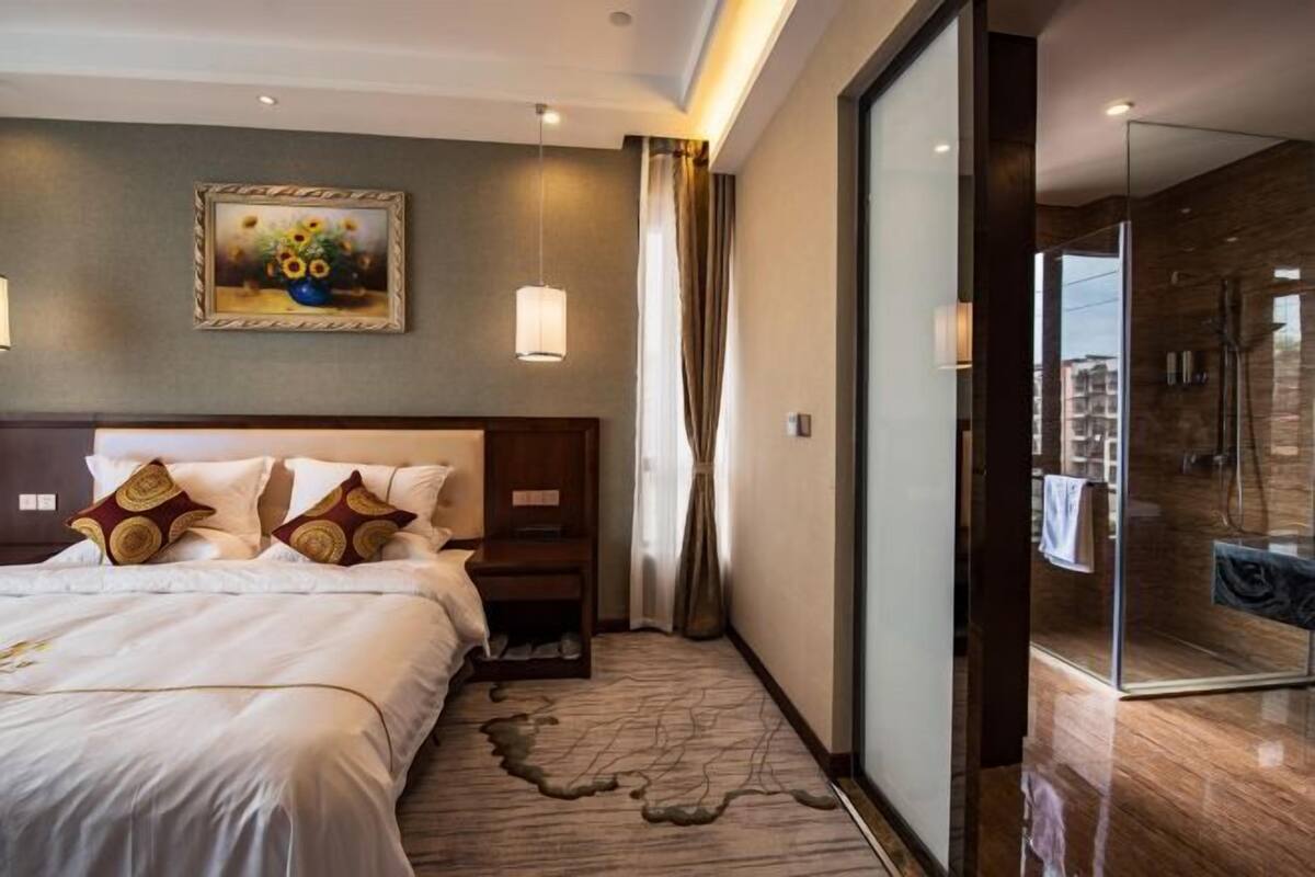A hotel room with a large bed, a painting on the wall, a glass shower area, and a view of the city.