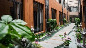Un pasillo moderno de hotel con paneles de madera, ventanas grandes y un camino de baldosas bordeado de plantas y flores en macetas.
