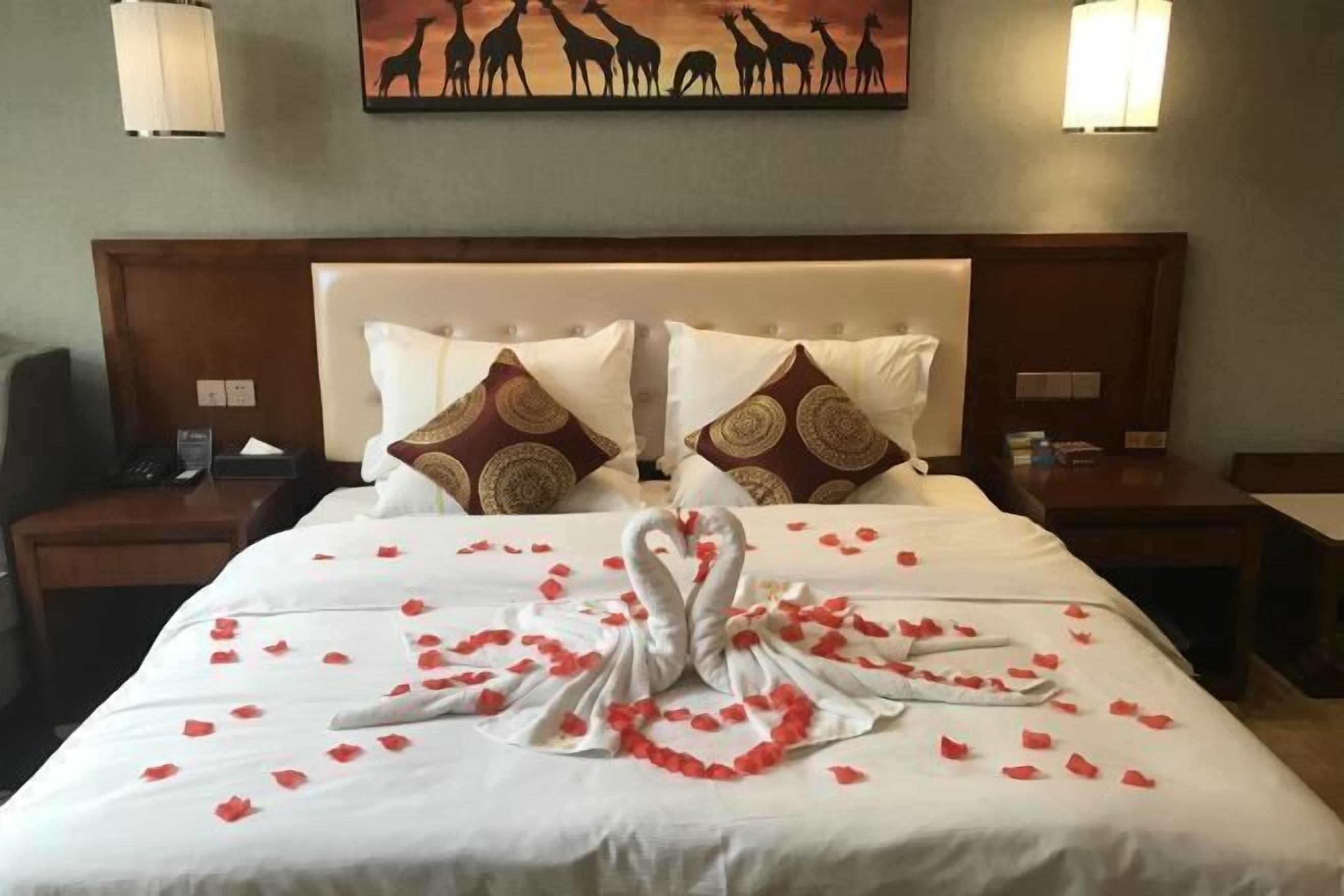 Una habitación de hotel con una cama grande, dos mesitas de noche, una obra de arte en la pared con jirafas y una decoración con forma de cisne hecha de pétalos de rosa.