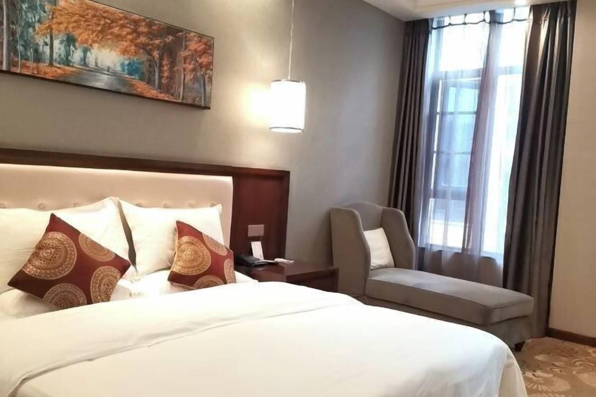 A hotel room with a large bed, a chair, a nightstand, a painting on the wall, and a window with curtains.