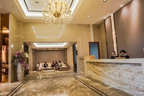 A hotel lobby with a marble reception desk, a chandelier, and a seating area with people.
