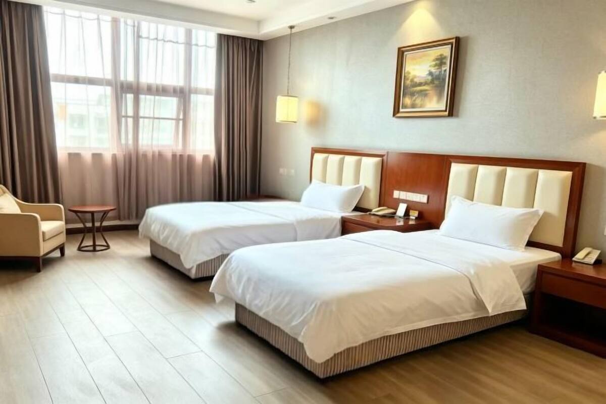 A hotel room with two beds, a chair, a small table, and a painting on the wall.