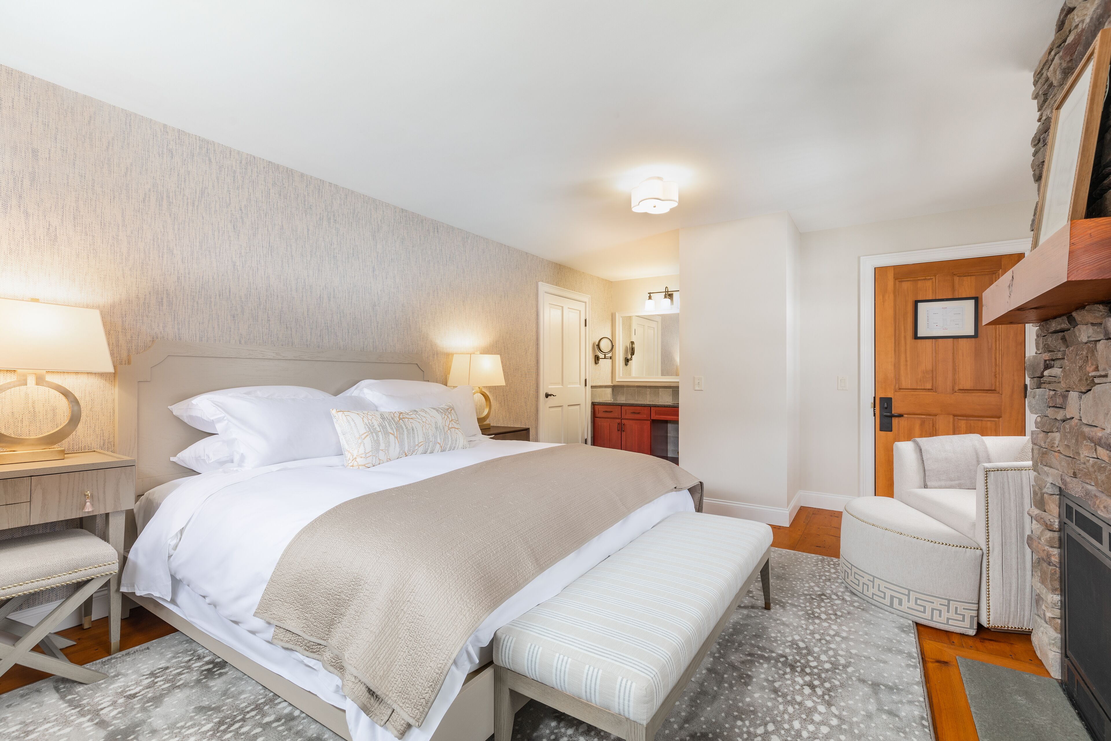 A bedroom with a bed, bedside tables, a bench, a chair, a fireplace, and a mirror.