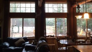 Interior acogedor de una cabaña con sofá de cuero, mesa de comedor de madera y ventanas amplias con vistas al bosque.