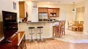 A modern kitchen with wooden cabinets, a stainless steel refrigerator, a built-in island with bar stools, and a dining area with wooden chairs.