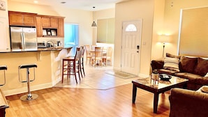 A spacious living area with a wooden floor, a brown sofa, a coffee table, and a kitchen area with wooden cabinets and a refrigerator.