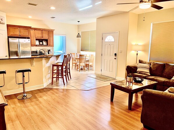 A spacious living area with a wooden floor, a brown sofa, a coffee table, and a kitchen area with wooden cabinets and a refrigerator.