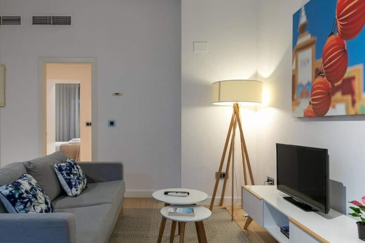 A modern living room with a grey sofa, a small round table, a floor lamp, and a flat-screen TV.