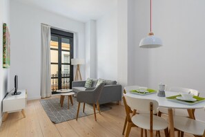 Ett modernt vardagsrum med en grå soffa, ett matbord med vita stolar, en golvlampa och ett fönster med utsikt över en balkong.