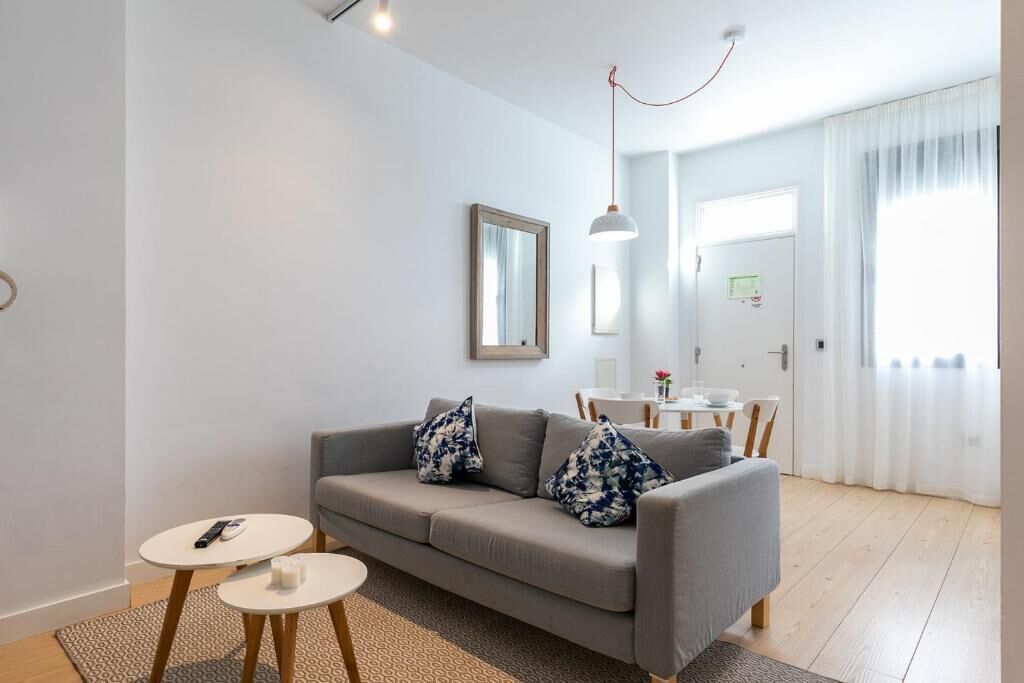 A modern living room with a grey sofa, two white round tables, a hanging pendant light, and a view of a dining area through a door.