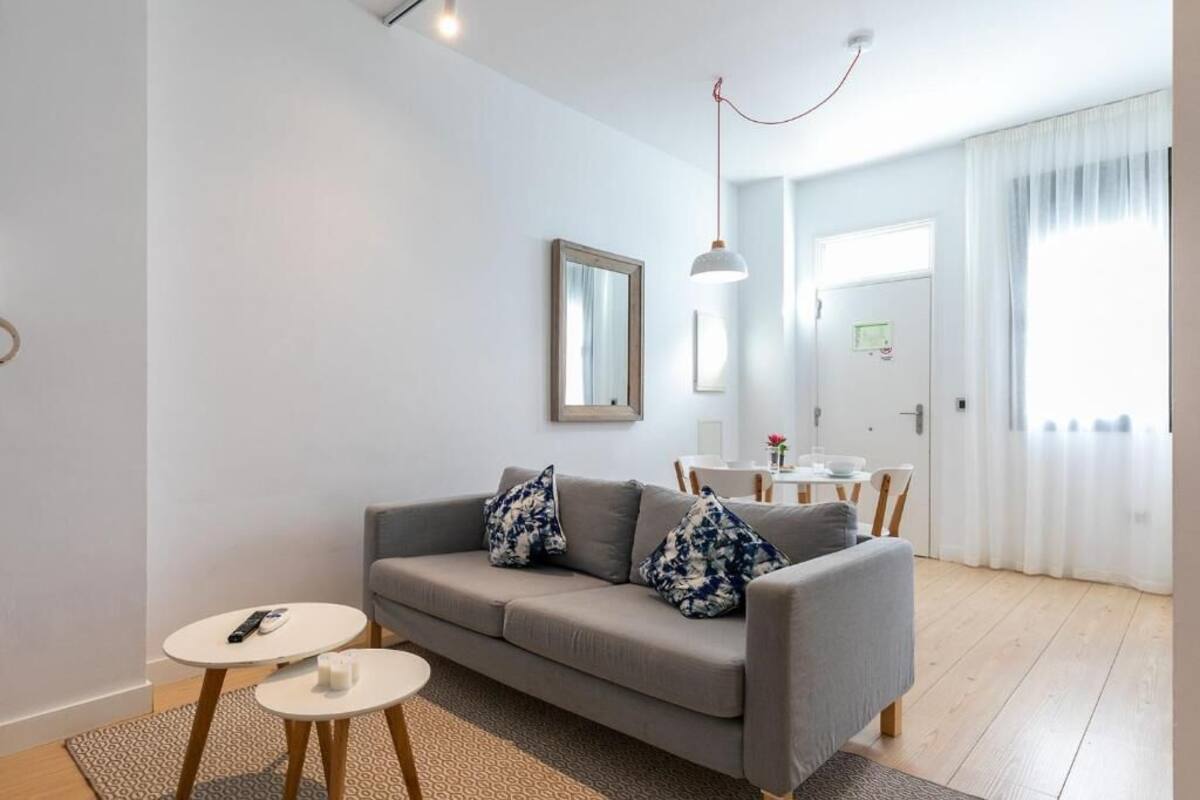 Un salon moderne avec un canapé gris, deux petites tables rondes blanches, un luminaire suspendu et une vue sur un coin repas accessible par une porte.