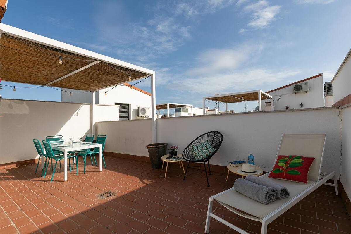 A rooftop terrace with a table and chairs, a lounge chair, and a small table with a hat and a decorative pillow.