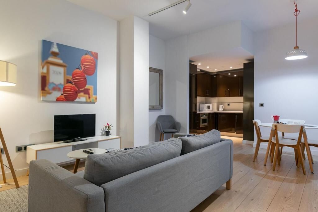 A modern living room with a grey sofa, a flat-screen TV, a dining area with a white table and chairs, and a kitchen visible through an open doorway.