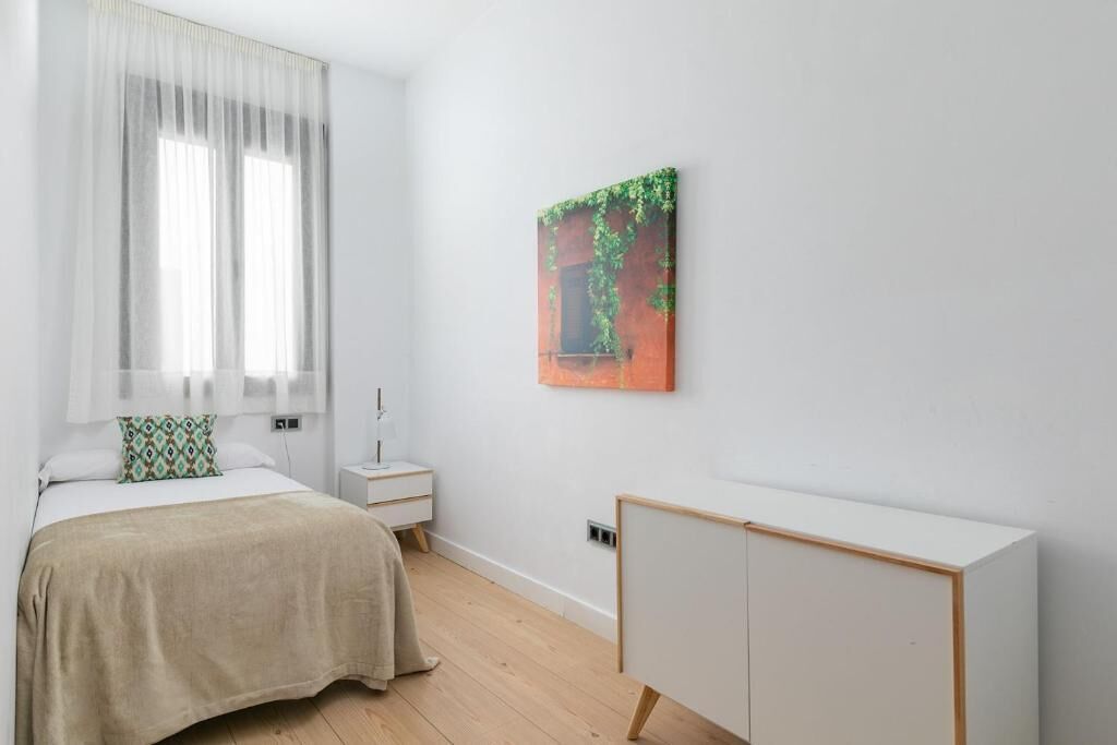 A bedroom with a bed, a nightstand, a bedside table, a wardrobe, and a window with sheer curtains.