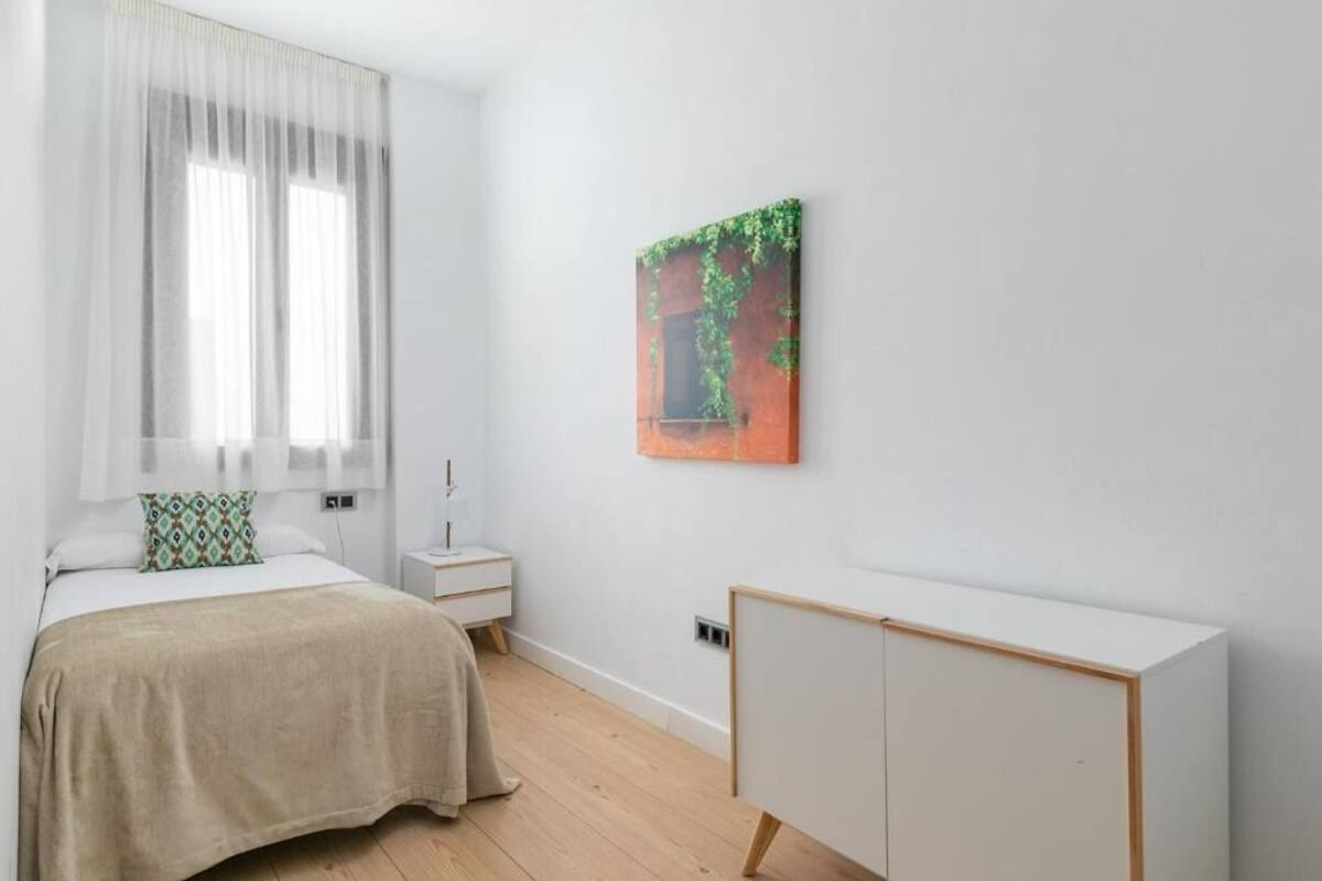 Une chambre à coucher comprenant un lit, une table de chevet, une commode, une armoire et une fenêtre avec des rideaux légers.