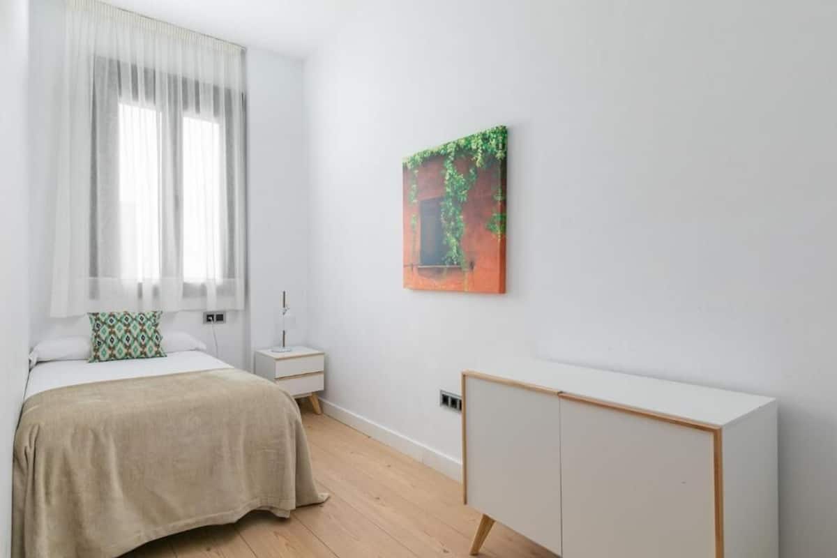 A bedroom with a bed, a nightstand, a bedside table, a wardrobe, and a window with sheer curtains.