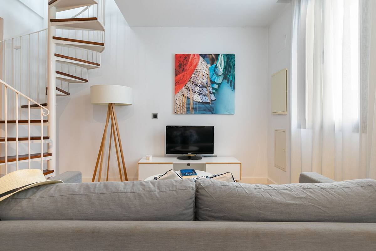 Un salon moderne avec un canapé gris, un téléviseur à écran plat posé sur un meuble blanc, un tableau accroché au mur et un escalier avec des rampes en bois.