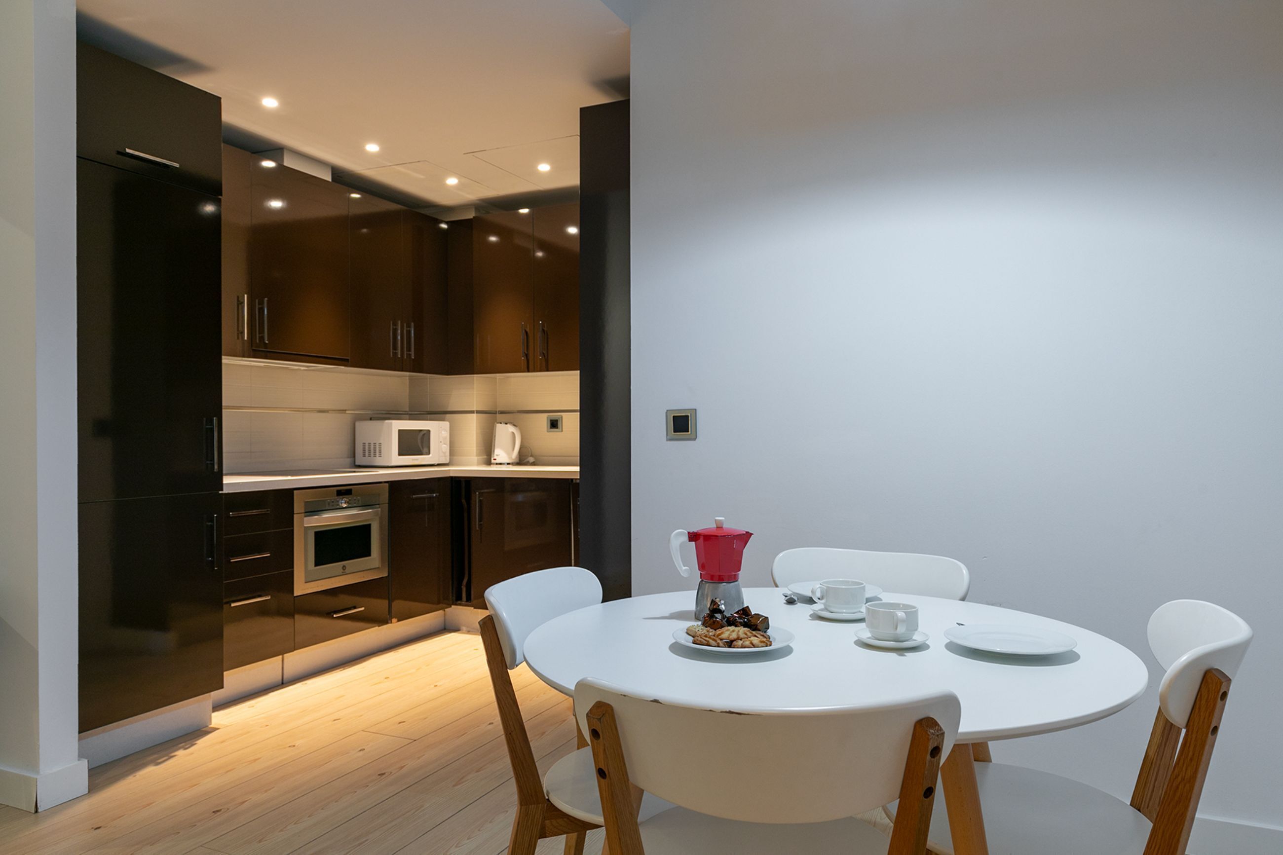 A modern kitchen with dark cabinets, a microwave, and an oven. There is a dining table with chairs and a coffee pot.