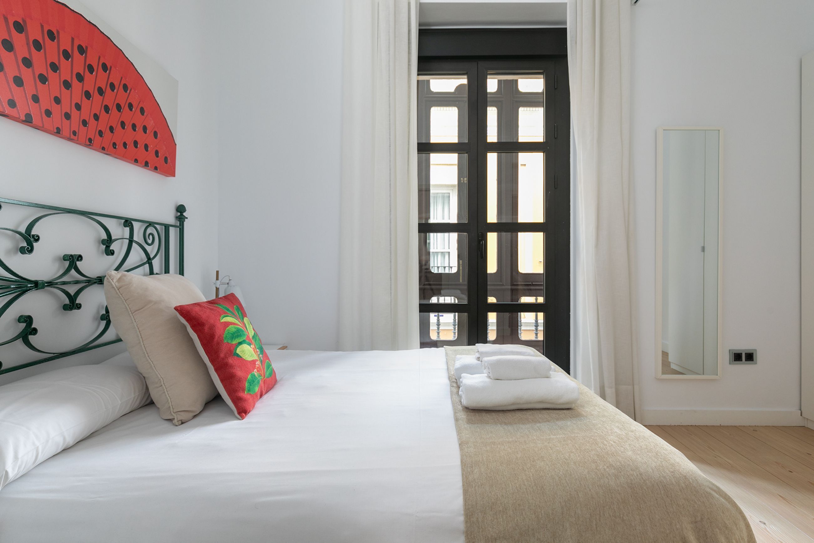 A hotel room with a bed, a red and green pillow, a white blanket, a mirror, and a view of the outside through a door.
