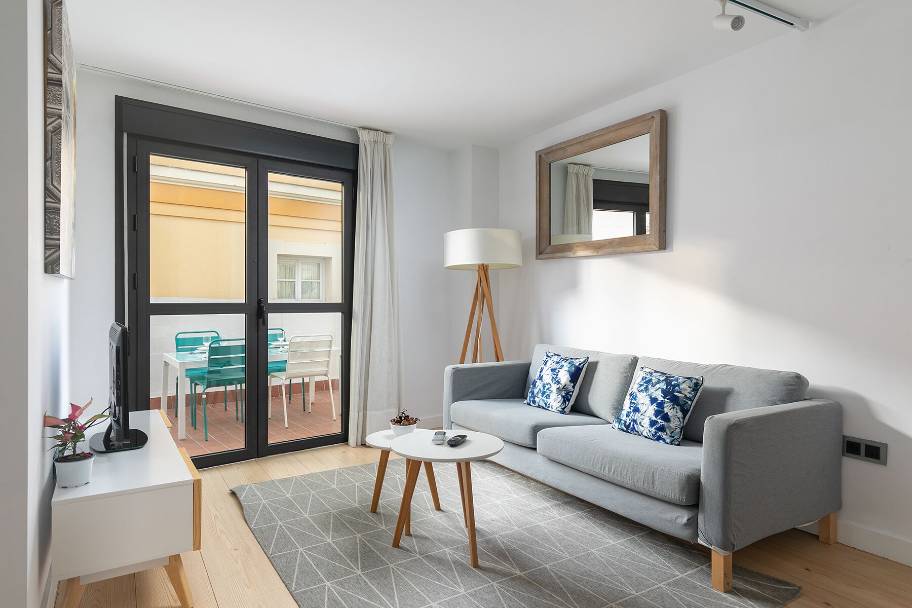 A modern living room with a grey sofa, two patterned cushions, a white coffee table, a floor lamp, and a sliding glass door leading to an outdoor area with green chairs and a table.