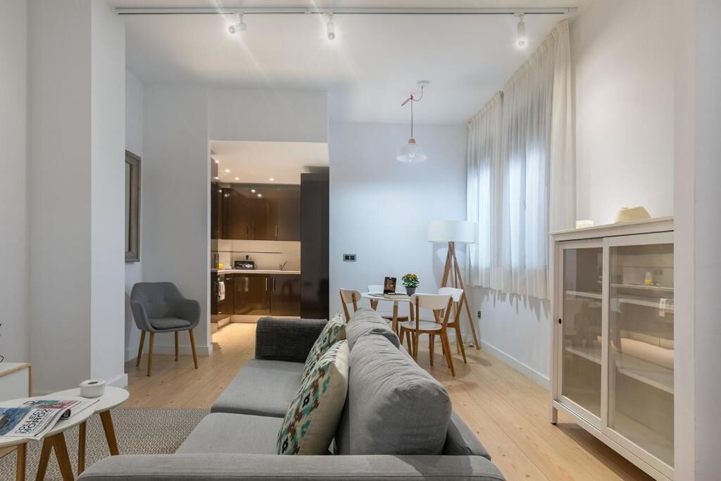 A modern living room with a grey sofa, a dining table with chairs, a kitchen area, and a cabinet with glass doors.