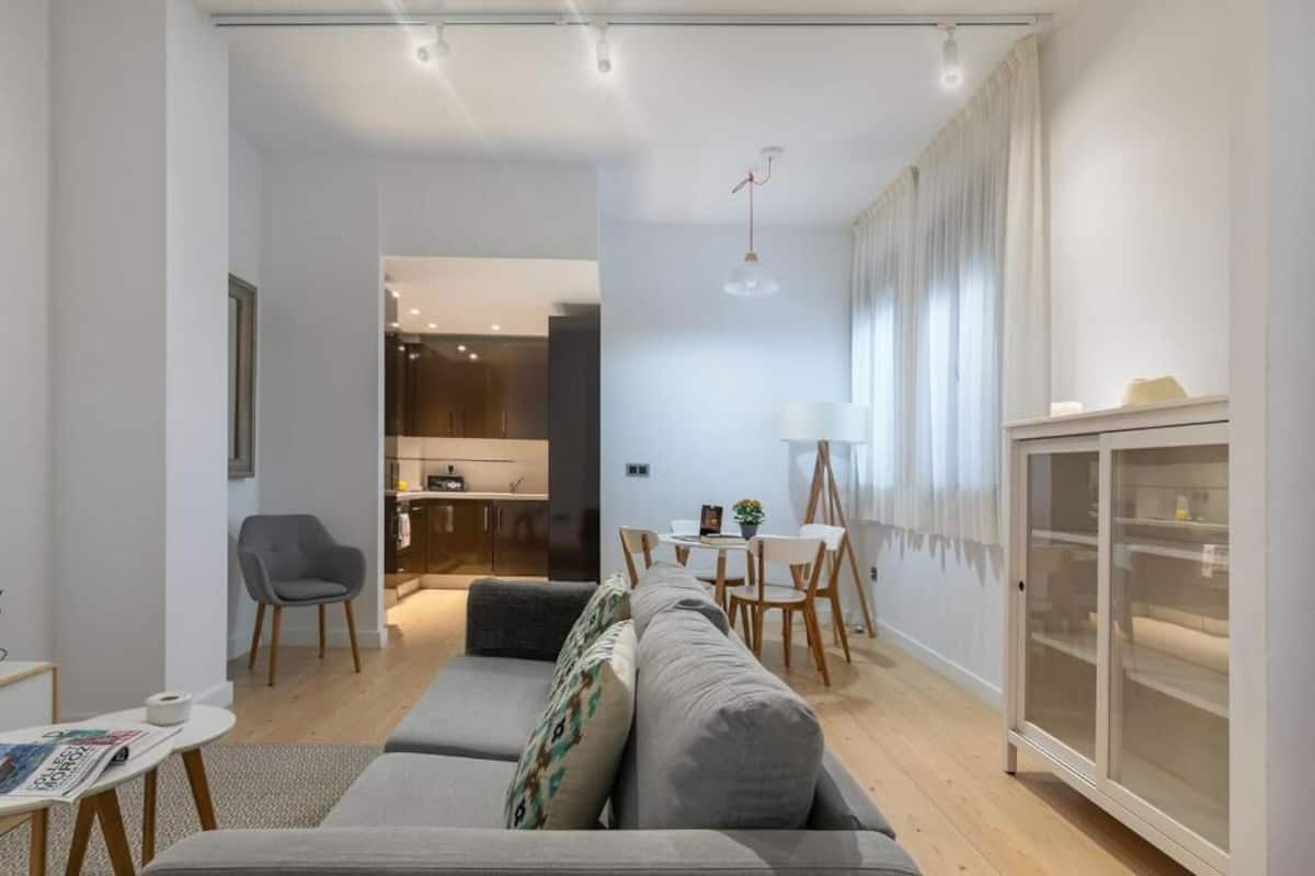 A modern living room with a grey sofa, a dining table with chairs, a kitchen area, and a cabinet with glass doors.