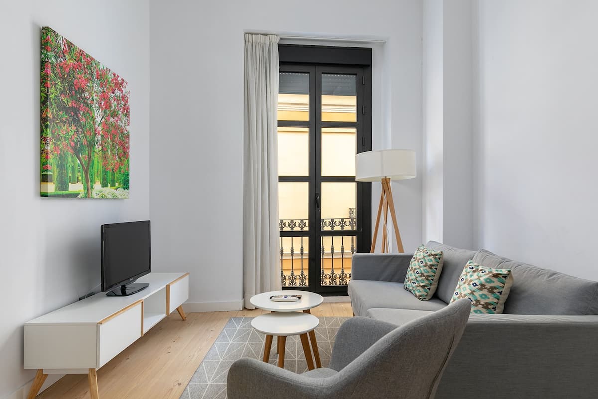 Un salon moderne avec un téléviseur à écran plat, un canapé gris, une petite table ronde et un tableau représentant un arbre.
