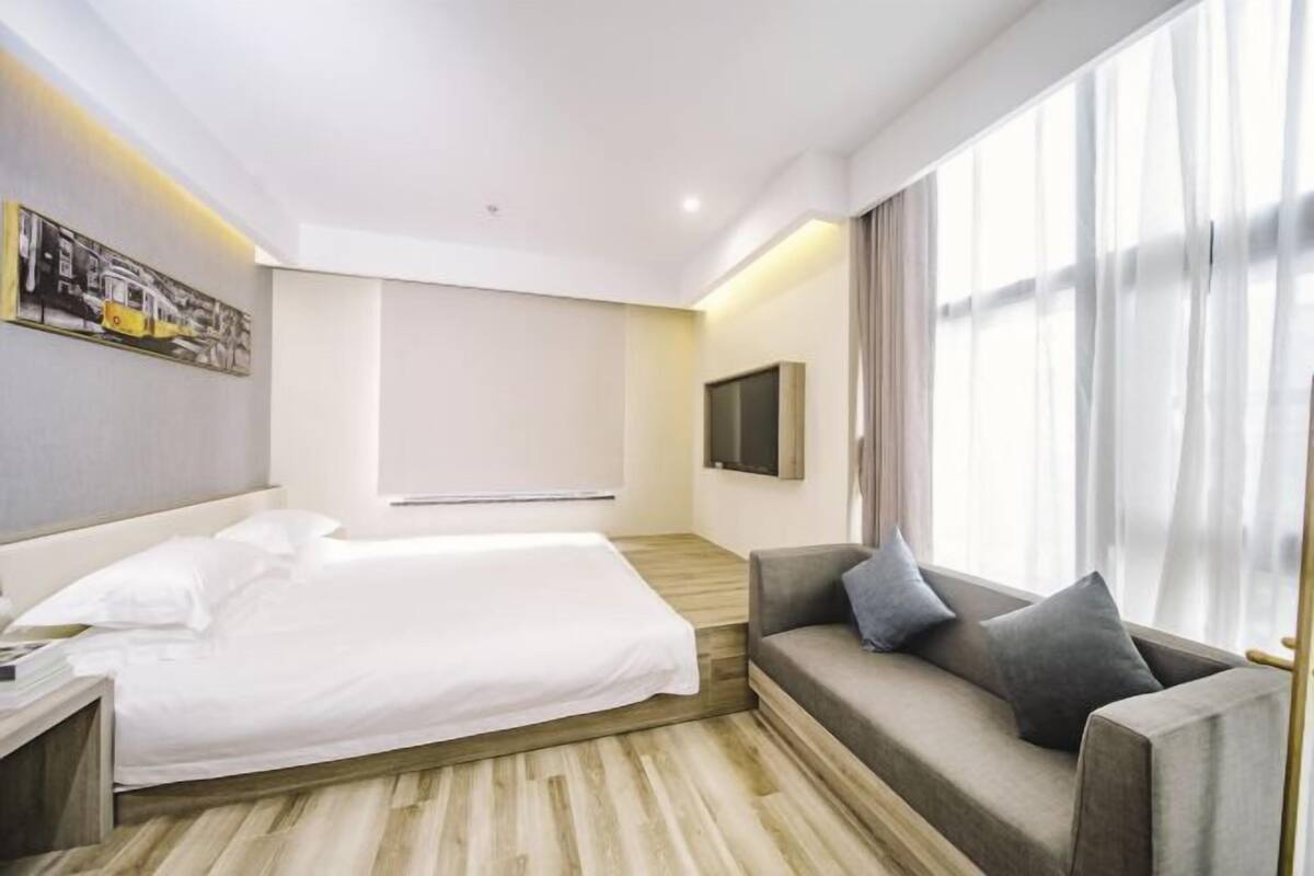 A hotel room with a bed, a sofa, a flat-screen TV, and a large window with curtains.