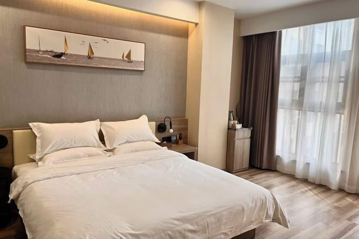 A hotel room with a large bed, a nightstand with a lamp, a painting of sailboats, and a view of the outdoors through sheer curtains.