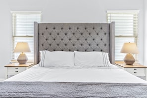 Cama bem arrumada com cabeceira estofada, roupa de cama branca e duas luminárias de mesa em mesas de cabeceira combinando.