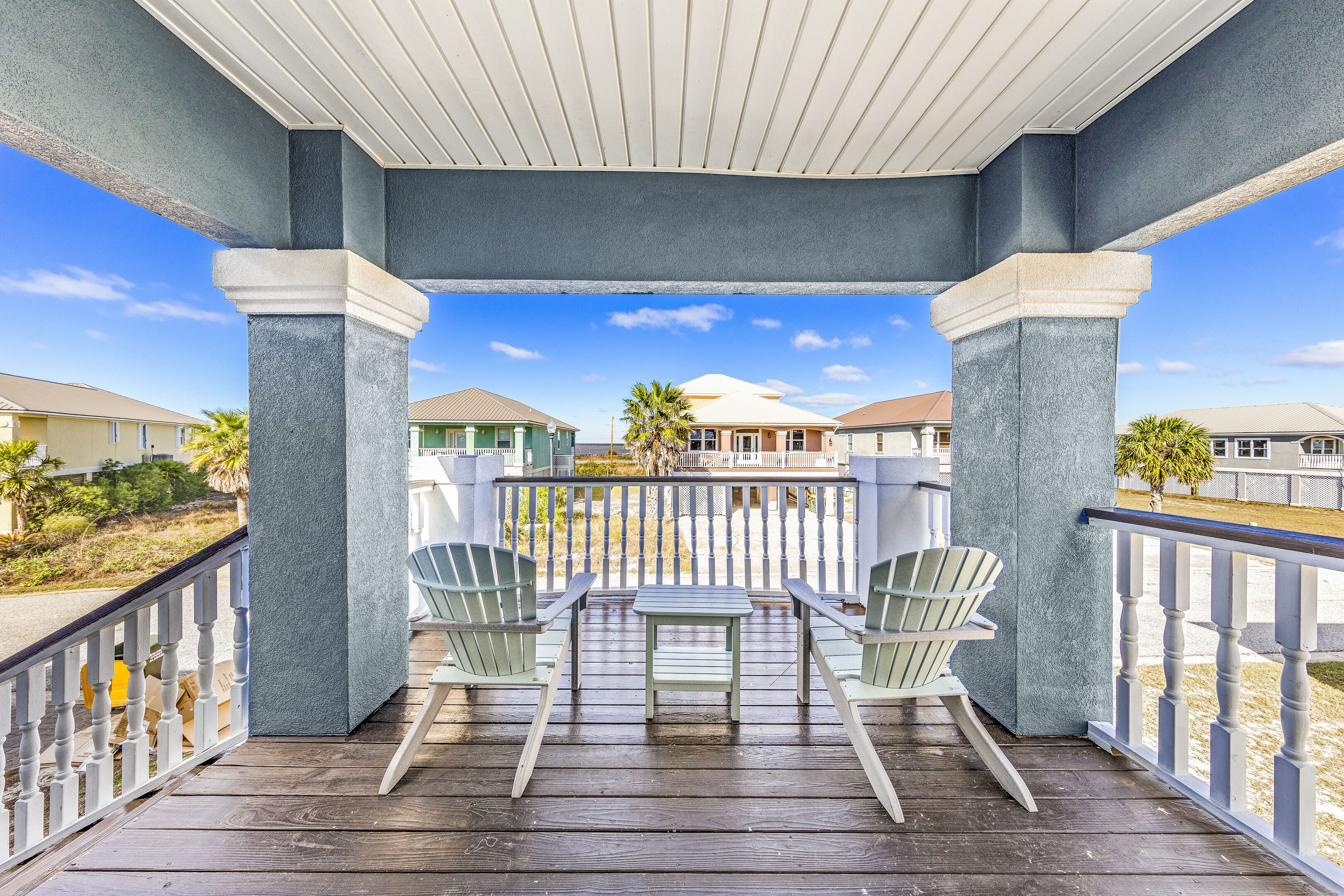 A covered porch with two Adirondack chairs and a small table, overlooking a residential area with houses and a palm tree.