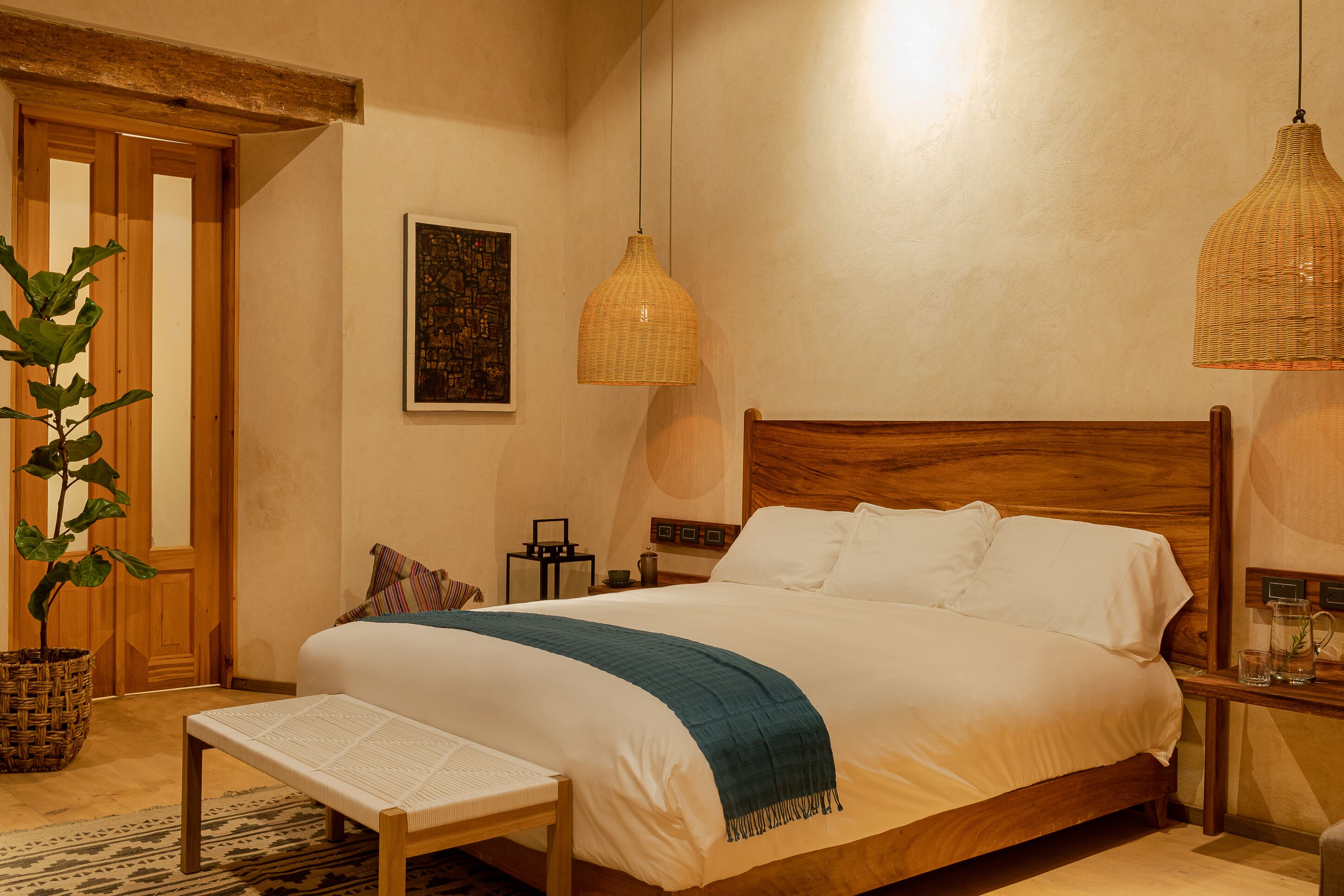A neatly made bed with a blue throw, a wooden headboard, and a hanging wicker lamp.