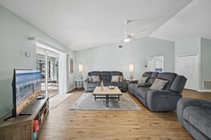 A spacious living room with a large flat-screen TV, grey sofas, a wooden coffee table, and a ceiling fan.