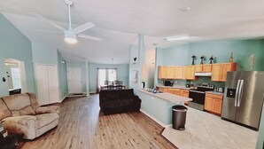 A spacious living area with a dark-colored sofa, a wooden floor, and a ceiling fan. A kitchen with wooden cabinets and stainless steel appliances is visible in the background.