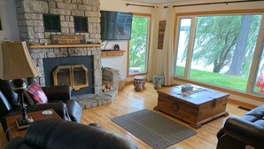 A cozy living room with a stone fireplace, leather sofas, a wooden coffee table, and large windows offering a view of the outdoors.