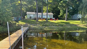 A wooden dock extending into a calm pond, surrounded by lush greenery and a house in the background.