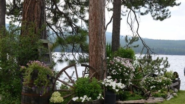 A rustic outdoor setting with a wooden wagon wheel, potted plants, and trees by a lake. - Lakefront home on McGregor Lake<br>Marion, Montana  (Marion)
