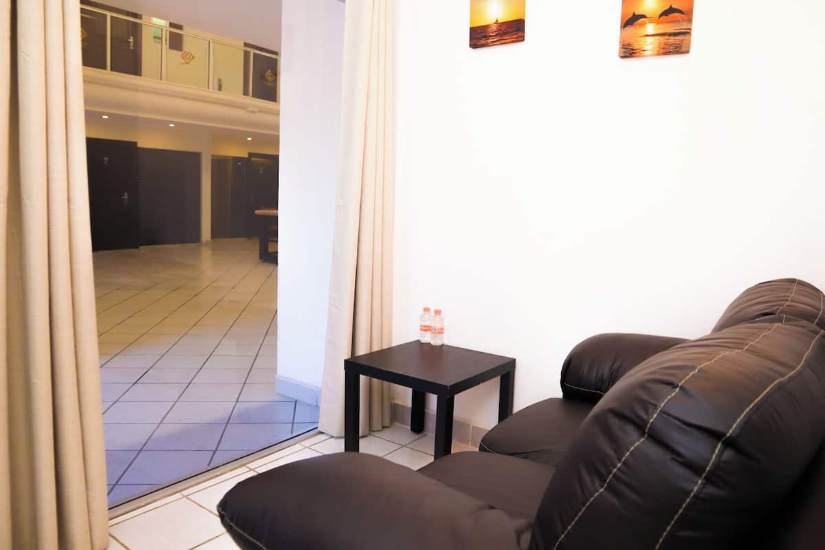 A room with a brown sofa, a small table with a bottle, and a view of a balcony with a door.
