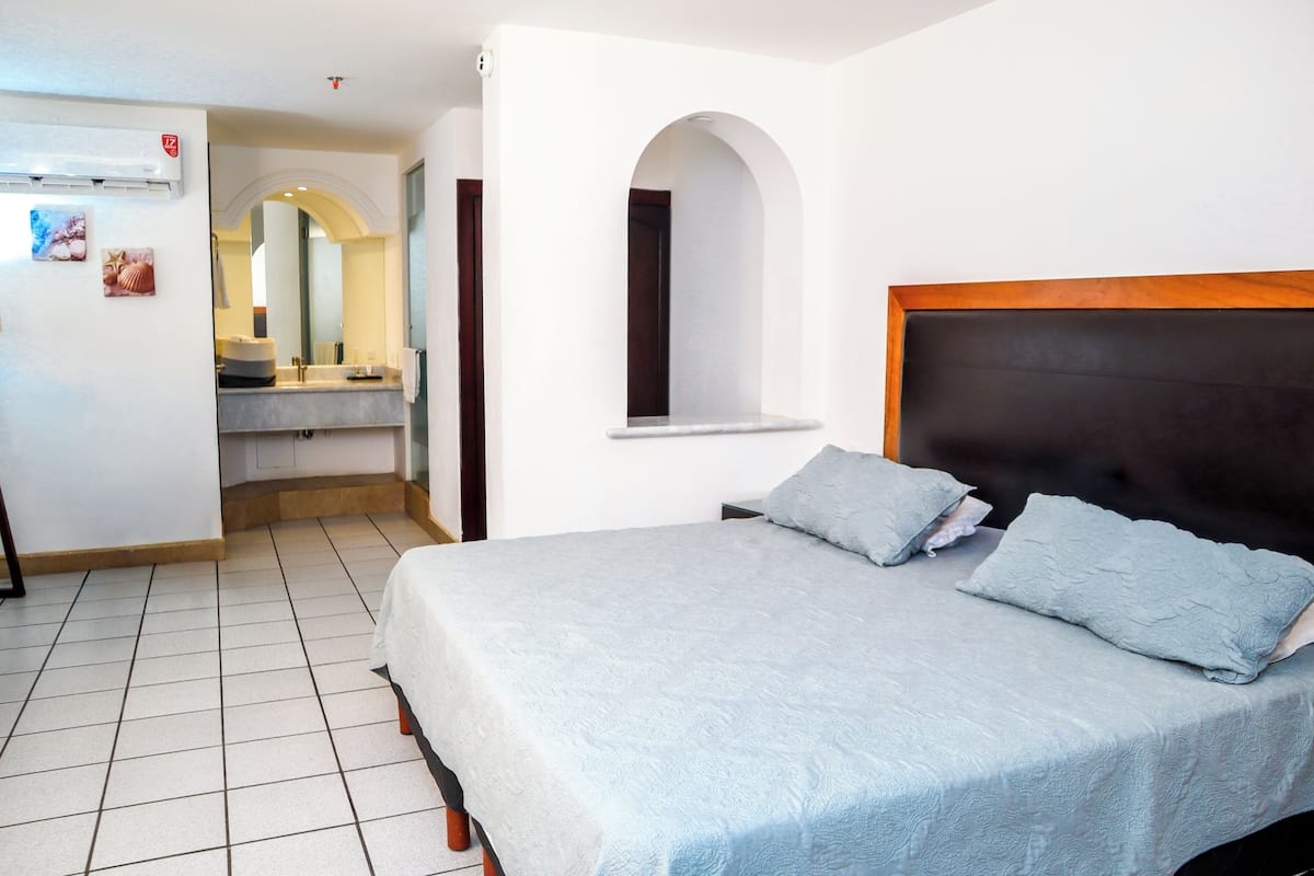 A bedroom with a bed, a window, a mirror, and a bathroom visible in the background.