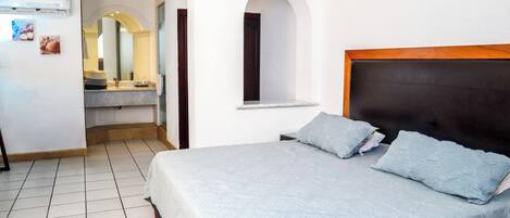 A bedroom with a bed, a window, a mirror, and a bathroom visible in the background.