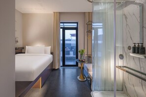 A hotel room with a bed, a small table with a vase, a shower area, and a view through a glass door.