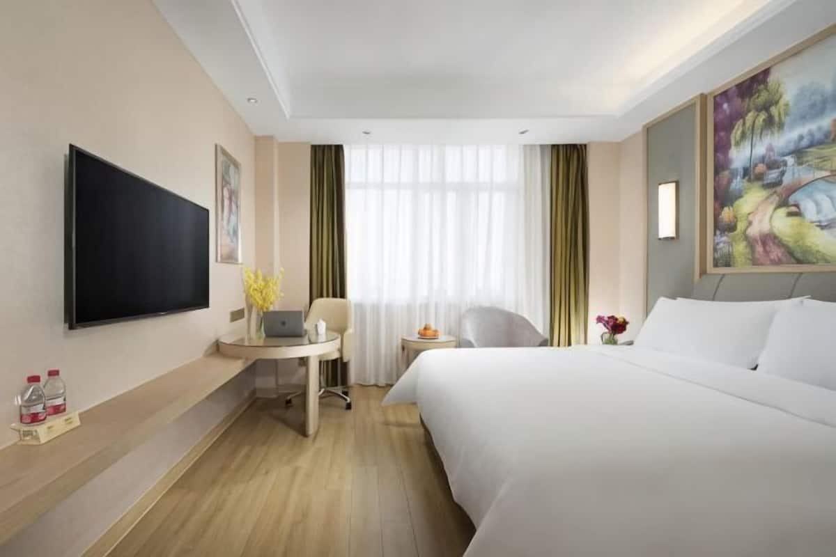 A hotel room with a bed, a desk with a laptop, a TV, and a painting on the wall.