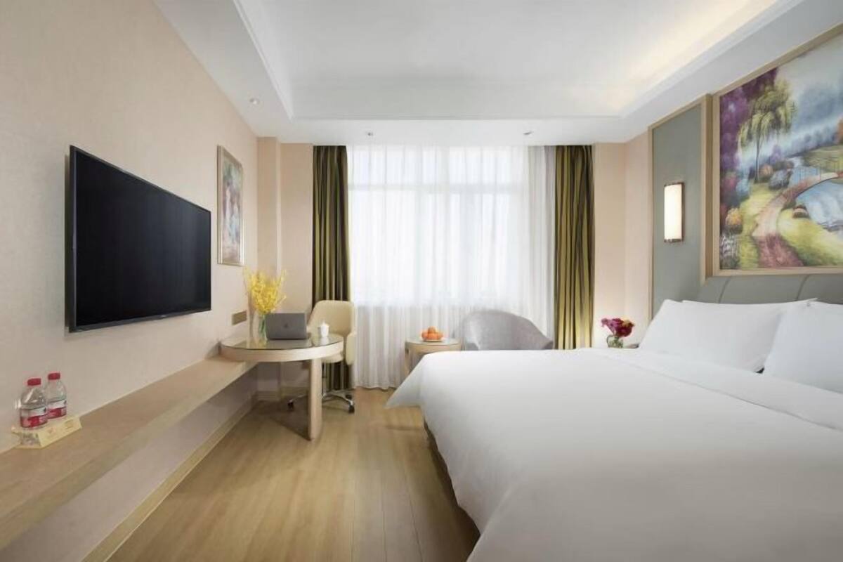 A hotel room with a bed, a desk with a laptop, a TV, and a painting on the wall.