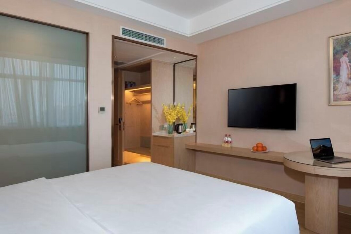 A hotel room with a bed, a TV, a desk with a laptop, a mirror, and a bathroom visible through an open door.