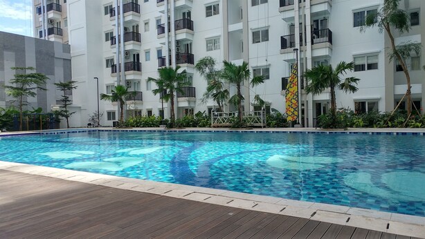 A swimming pool with a wooden deck, surrounded by lush greenery and a multi-story building with balconies. - Cozy and Modern Signature Park Grande Studio Apartment (Jakarta)